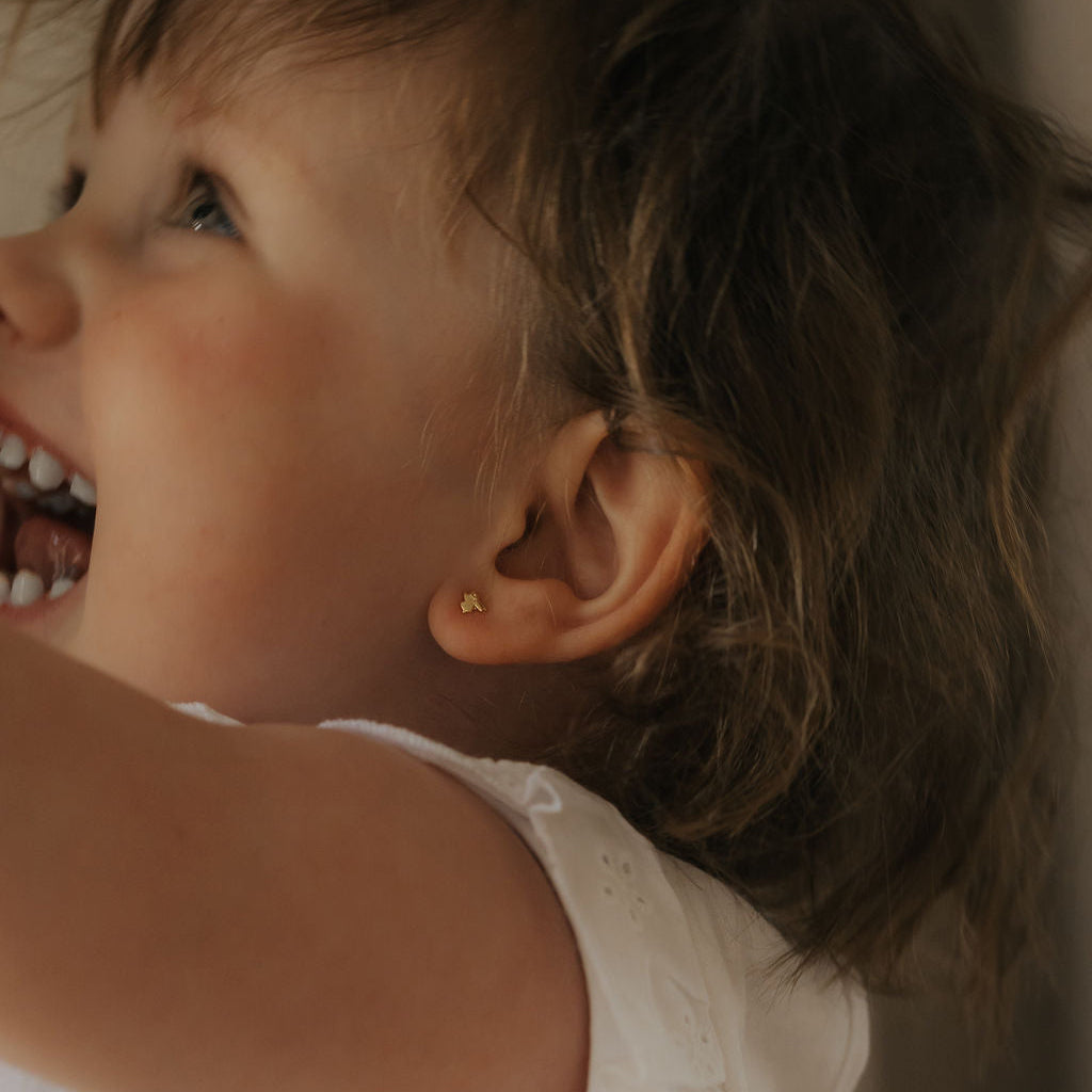 A close-up lifestyle shot of a young child wearing a tiny gold stud earring, looking upward and smiling. The child is wearing a white sleeveless top, set against a soft, warm beige background.