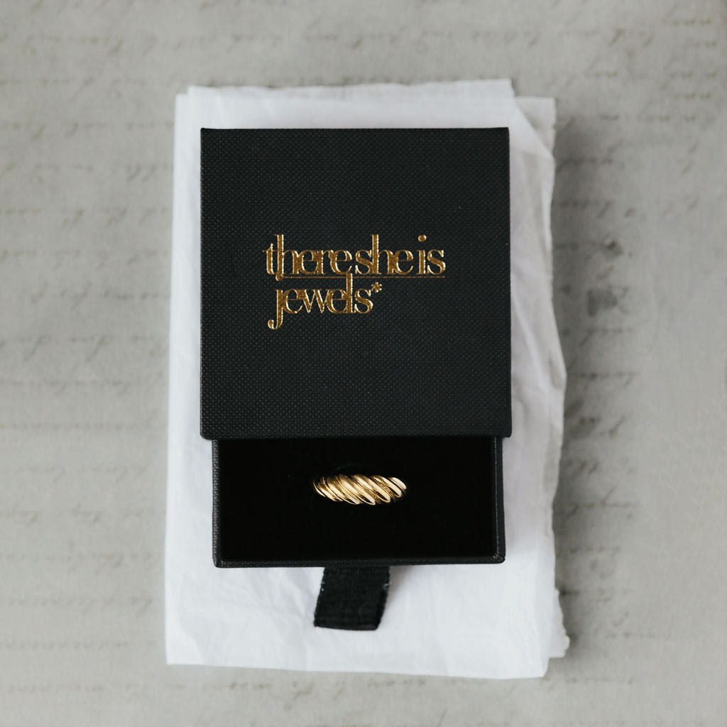Gold ring in a black box with 'theresheisjewels' branding on a textured surface
