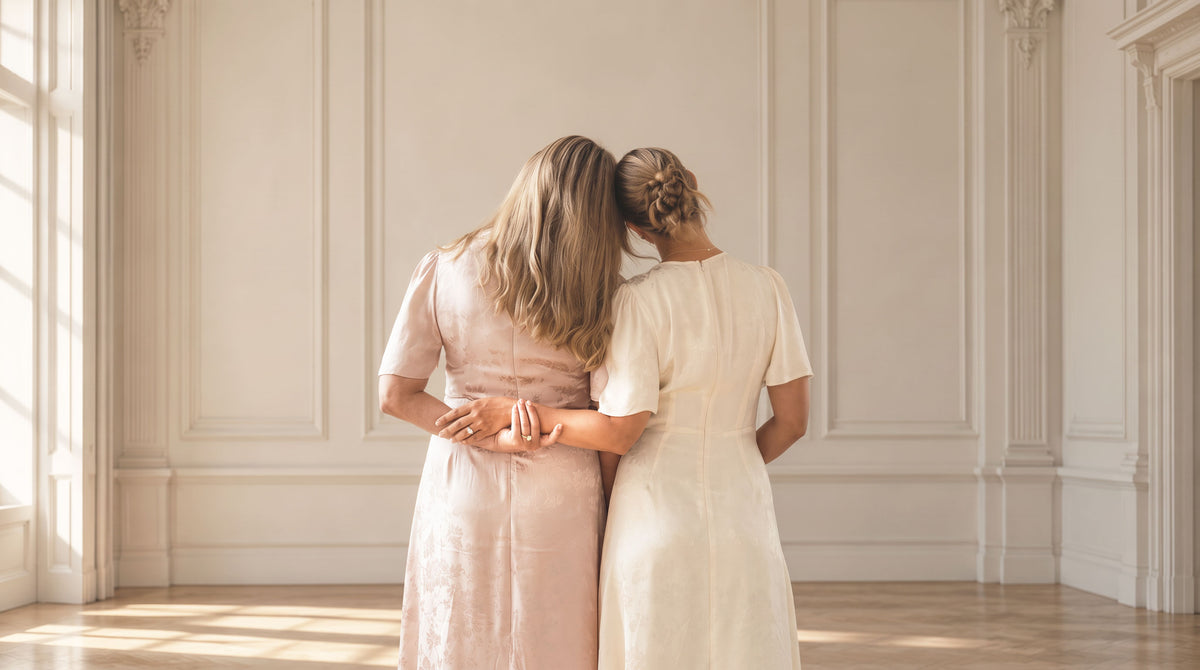 Two women standing with their backs to the camera in a bright, elegant room with cream walls and wood floors, wearing simple midi dresses in pale pink and cream.